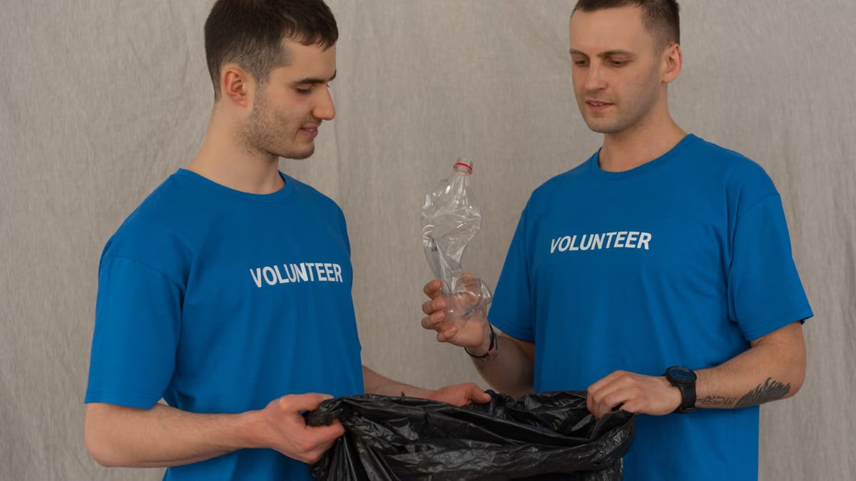 Two male volunteers in blue shirts collecting plastic bottles for recycling indoors.